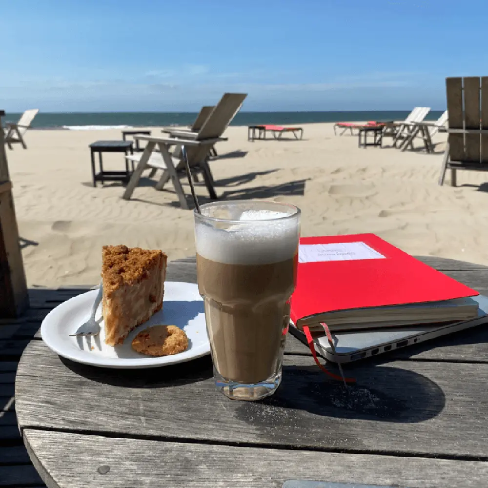 Ein Latte Macchiato sowie ein Teller mit einem Stück Kuchen und einem Keks neben einem roten Notzbuch, das auf einem Laptop liegt auf einem Tisch am Strand in Den Haag als Teil von "12 von 12".