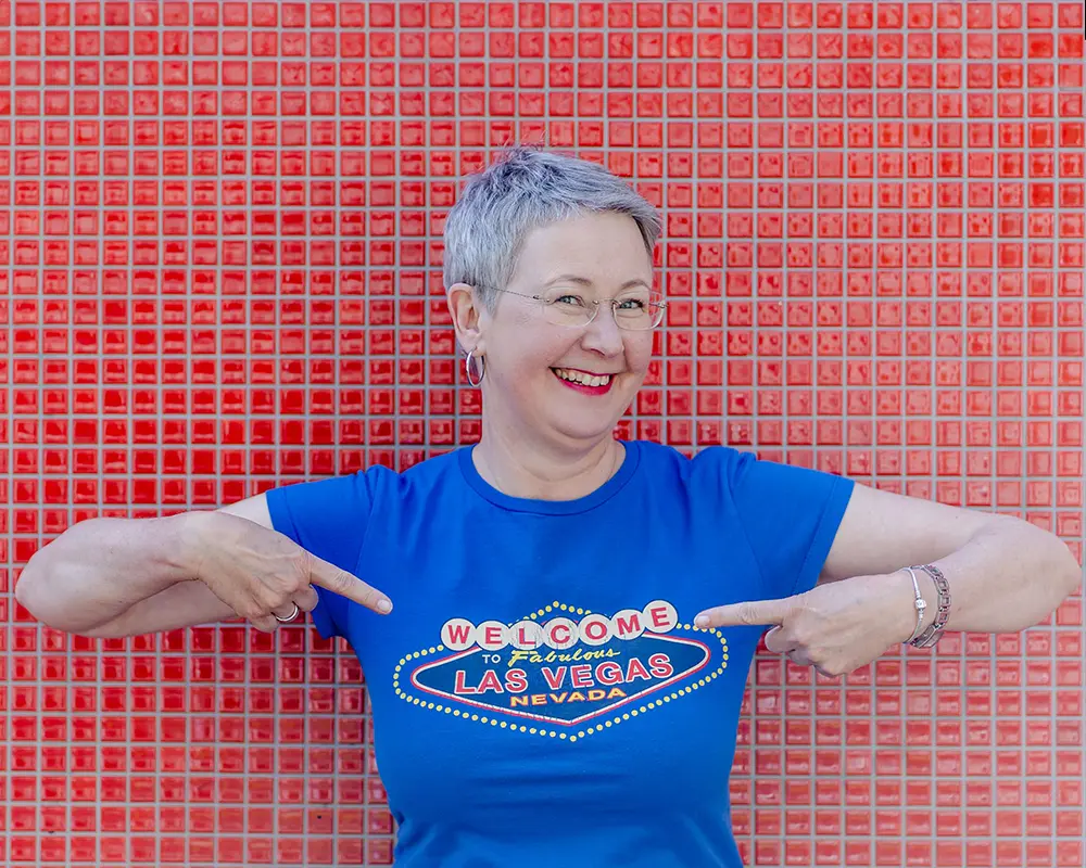 Marketing-Mentorin Susanne Jestel steht im knallblauen T-Shirt vor einer rot gefließten Wand und zeigt lachend mit beiden Zeigefingern auf sich selbst als Symbolbild worauf sie sich freut.
