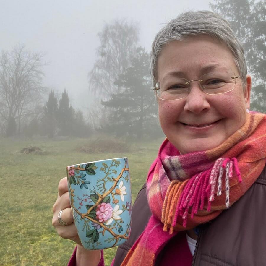 Susanne Jestel steht anlässlich von "12 von 12" im nebligen Garten des Eichhof Cottages an der ostdeutschen Küste und blickt lächelnd in die Kamera. Sie trägt einen karierten Schal und hält in der rechten Hand eine bunt geblümte Tasse.