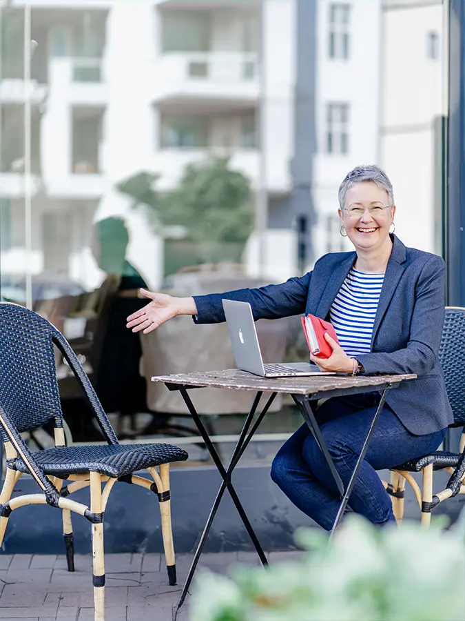 Marketing-Mentorin Susanne Jestel sitzt mit ihrem Laptop und einem roten Notizbuch an einem Tisch im Straßencafé in Berlin, streckt lächelnd die Hand aus und lädt ein, bei ihr Platz zu nehmen.