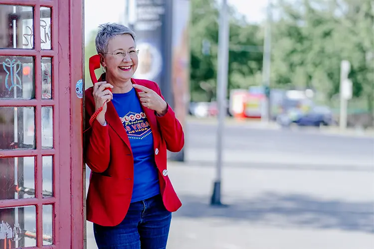 Susanne Jestel steht in einem roten Blazer an einer roten Telefonzelle und hält einen roten Telefonhörer ans Ohr auf den sie lächenld mit den anderen Hand zeigt als Symbolbild, um Kontakt mit ihr aufzunehmen und ins Gespräch zu kommen.