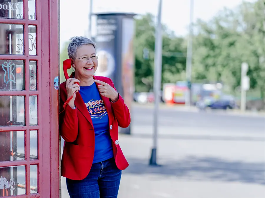 Susanne Jestel steht in einem roten Blazer an einer roten Telefonzelle und hält einen roten Telefonhörer ans Ohr auf den sie lächenld mit den anderen Hand zeigt als Symbolbild, um Kontakt mit ihr aufzunehmen und ins Gespräch zu kommen.