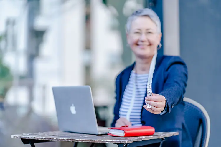 Susanne Jestel sitzt hinter ihrem Laptop und einem roten Notizbuch an einem Bistrotisch und hält ein Maßband in die Kamera als Symbolbild für die Seite Über mich und ihre Arbeit mit kleinen Kampagnen und dem Marketing Atelier.