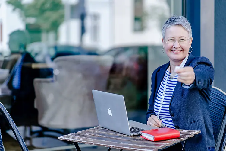Susanne Jestel sitzt lächend an ihrem Laptop in einem Straßencafé und hält ein Maßband in die Kamera als Symbolbild für die Zusammenarbeit mit ihr im Marketing Atelier.
