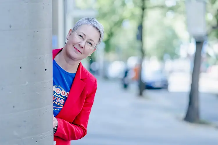 Susanne Jestel schaut in einem roten Blazer hinter einer Betonsäule hervor als Symbolbild für die Zusammenarbeit mit ihr.