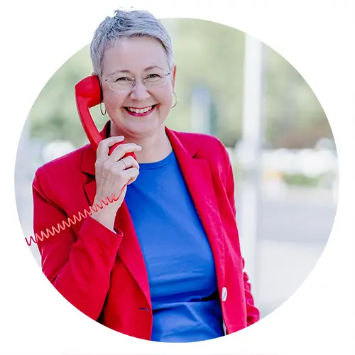 Susanne Jestel hält sich einen roten Telefonhörer ans Ohr und lächelt in die Kamera als Symbolbild für ein Kennenlerngespräch mit ihr.
